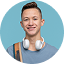 Smiling young man with short hair wearing a gray shirt and headphones around his neck on a blue background.